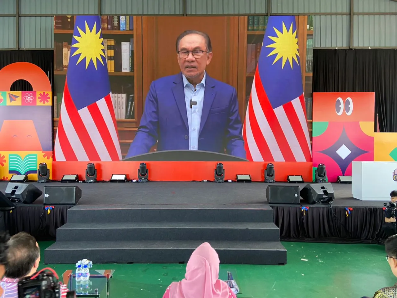 A speaker addressing the Program Anak Kita launch by video, between two Malaysian flags
