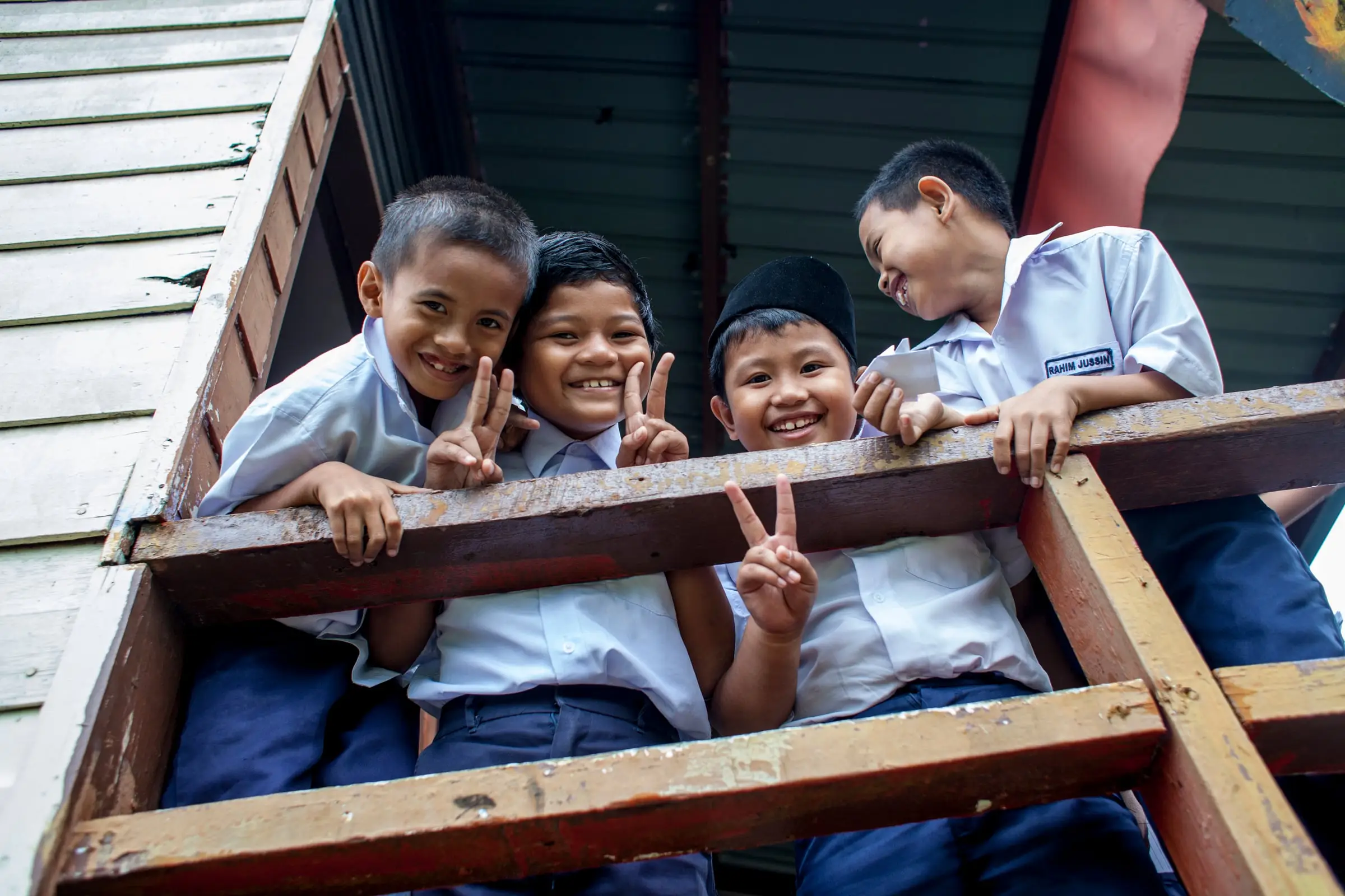 Four boys leaning out of a wooden school window frame, making peace signs, at SK Kimanis, Papar